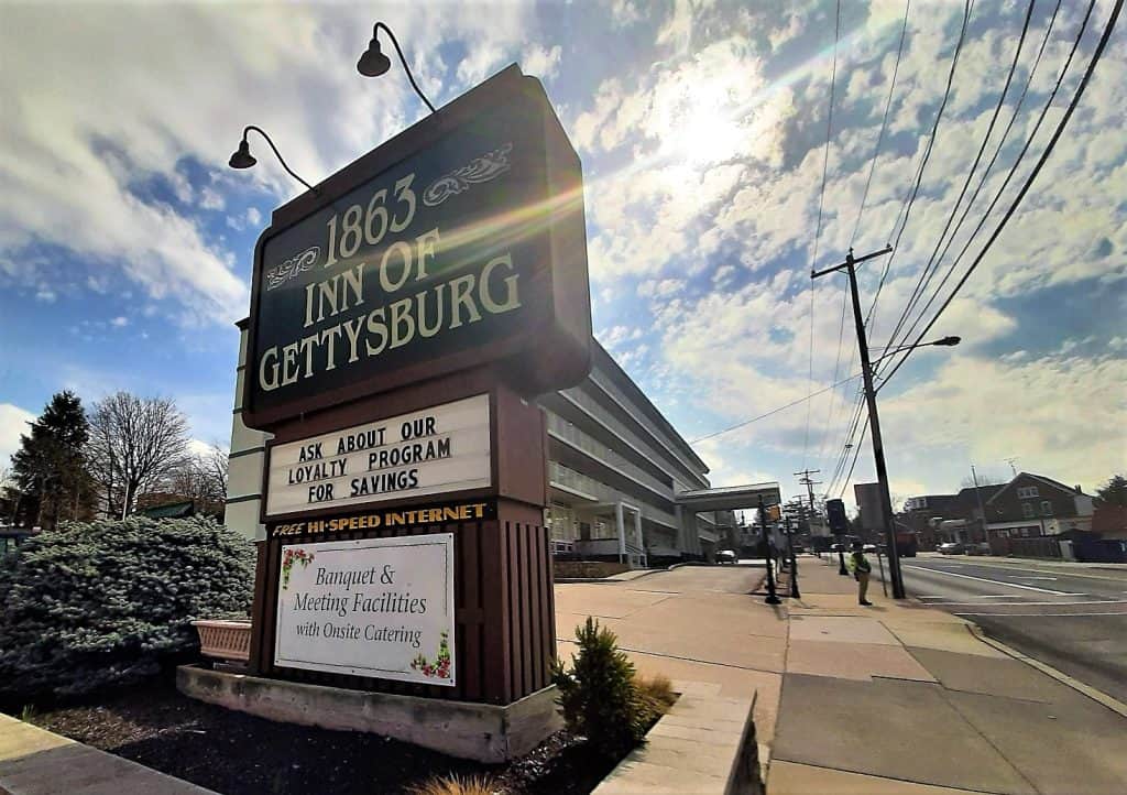 Green sign for 1863 Inn of Gettysburg with hotel in background against sunny and hazy sky.