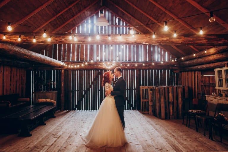A bride and groom kiss on the wooden dance floor of a barn with light coming in through slats and additional white lights hanging from rafters of a barn.