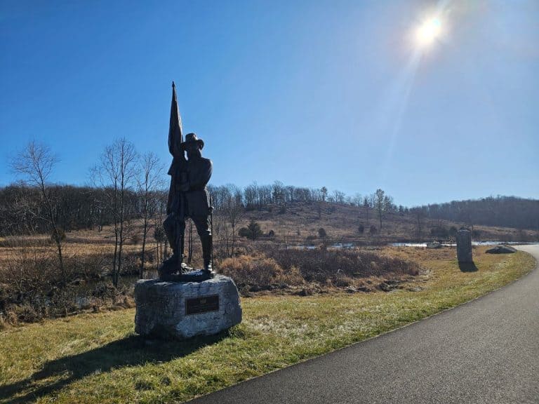 A statue of a soldier holding a flagpole with Gettysburg Little Round Top in the background under a blue sky and shining sun.