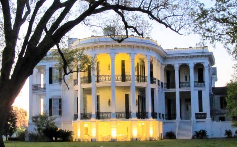 The oval front of Nottoway Plantation has lights shining at twilight. The building is white with large white pillars and black railings.