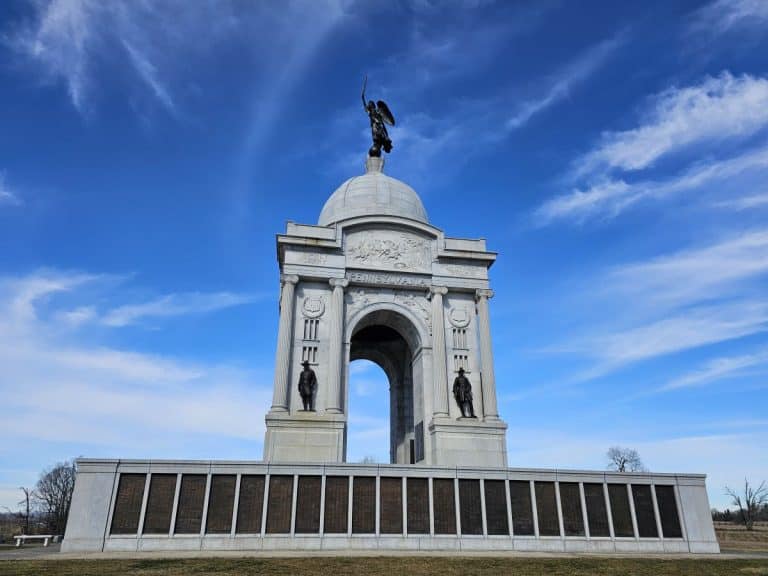 The Pennsylvania Monument, a must see Gettysburg monument because it is the largest on the battlefield. It is white with a dome top and an angel holding a torch on top, under a blue sky.