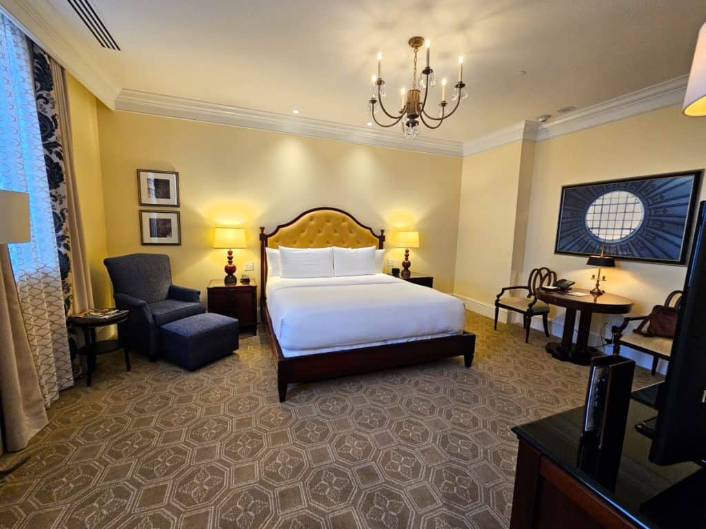 The bedroom at The Jefferson shows a bed with white coverings and gold headboard with a chandelier hanging above. A blue lounge chair is on the left and a desk on the right.