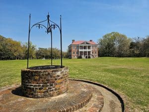 The stately Drayton Hall Plantation is a two-story red brick structure with a white two-story balcony in the center. It is in the background, with a red circular brick well in the foreground.