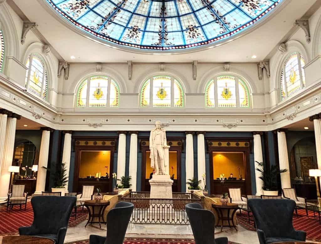 The white marble statue of Thomas Jefferson is part of the history of the Jefferson Hotel in Richmond VA. It stands beneath a colorful stained glass dome, with white columns in the background.