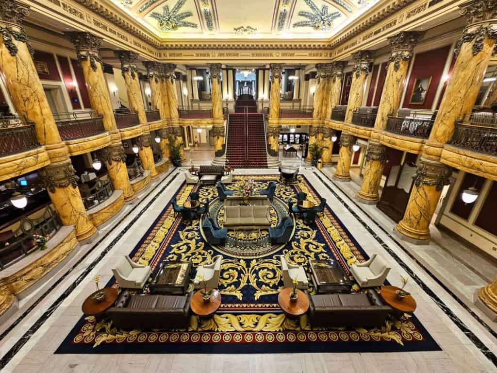 The magnificent lobby of The Jefferson Hotel in Richmond VA with large tan pillars draped in intricate vine and grape clusters. There is a sitting area with a blue and gold rug.