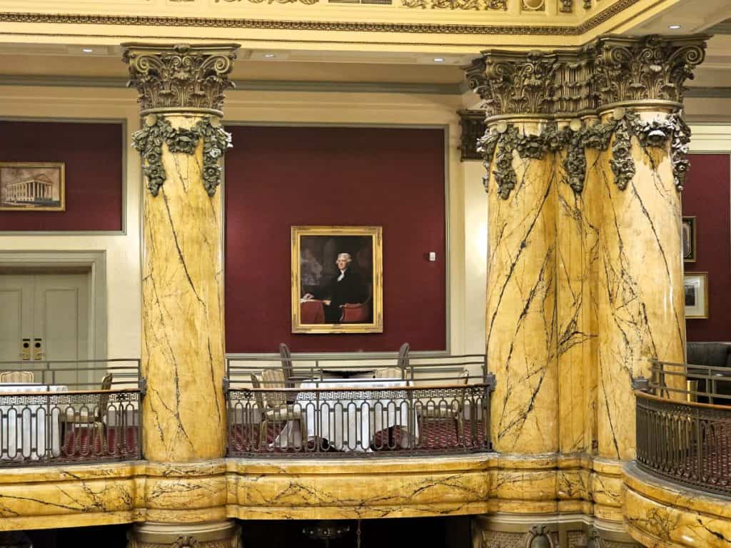 A beautiful reproduction oil painting hangs between two faux marble columns against a red wallpapered wall at The Jefferson Hotel.