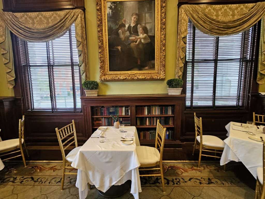 An original portrait in a gold gilded frame hangs above a bookcase, with a table with a white tablecloth in the foreground.