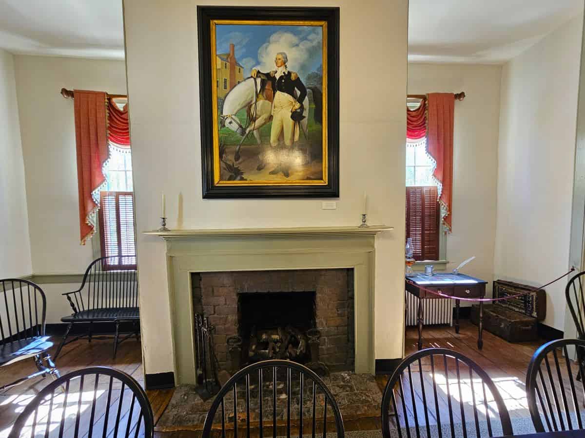 A painting of George Washington with a white horse is seen on the wall above a fireplace with period chairs in front.