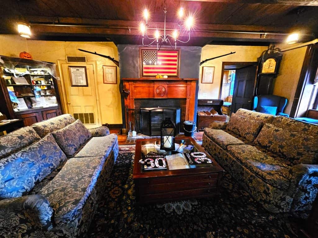 Two couches face each other with a fireplace in the background in the lobby of the Beekman Arms Inn. A chandelier hangs overhead and a flag hangs over the fireplace.