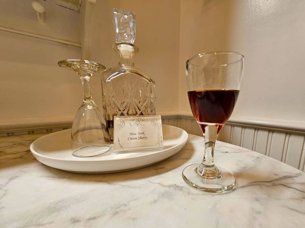 A glass of sherry sits in front of a decanter on a dresser top.