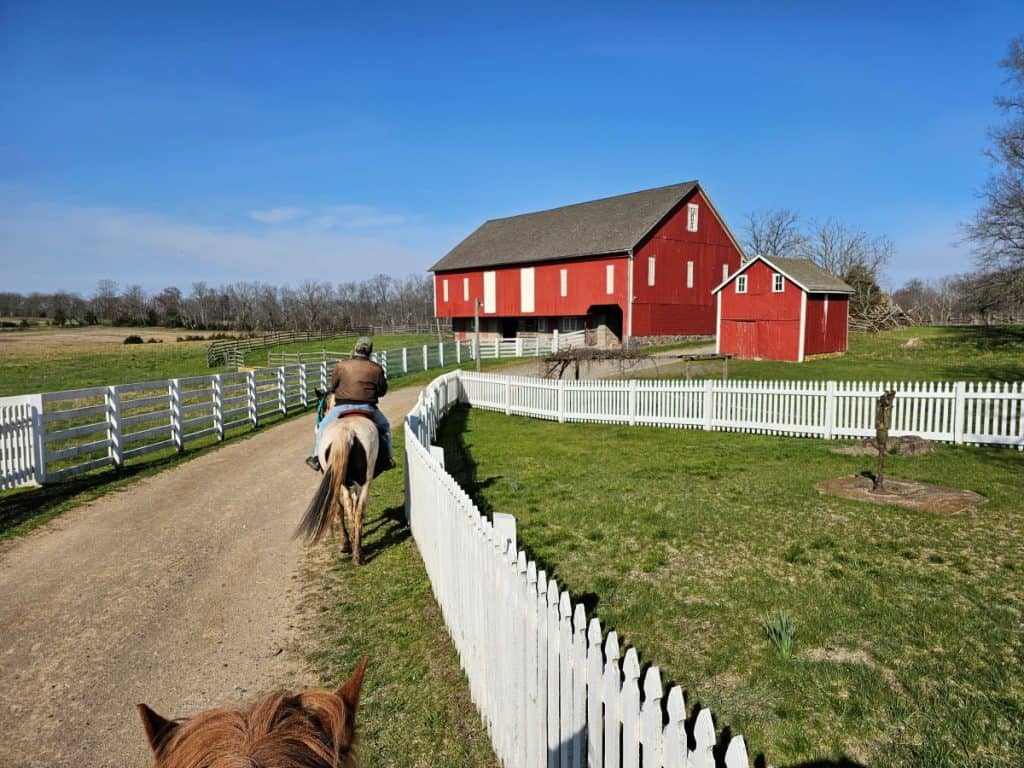 A horse and rider walk past a white picket fence and toward a big red barn. A horseback ride is a great way to see the Gettysburg battlefield in the spring.
