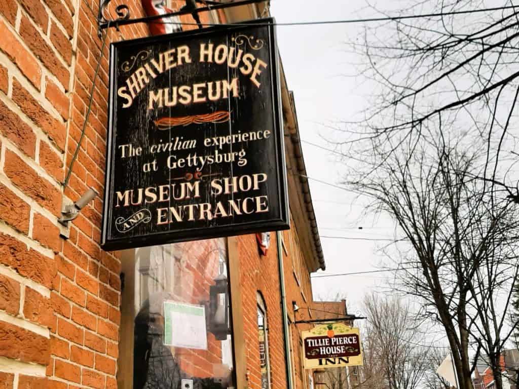 A sign that says Shriver House Museum is brown with cream lettering and hangs on the side of a brick building.