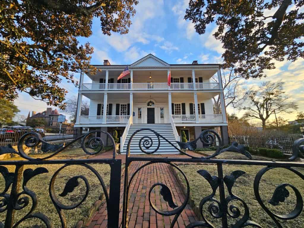 The Burgwyn-Wright Mansion is one of the must-see historic sites in Wilmington NC. It is a two story tall house, framed by tree limbs and with a wrought iron fence in front.