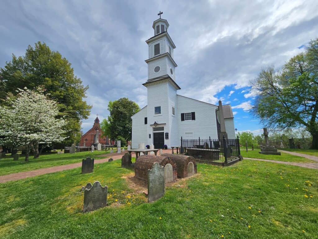 St. Johns Church is a white church against a cloudy sky, with a graveyard in front.