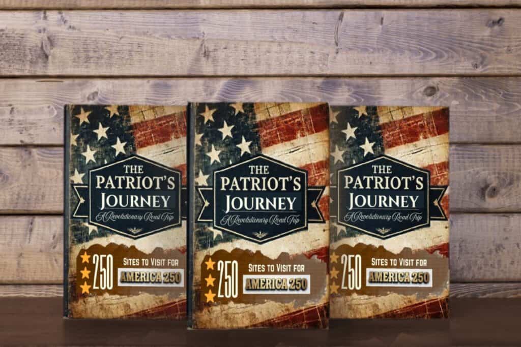 Three books with a flag on the front that says The Patriot's Journey against a wood wall. The book helps promote travel for America 250.